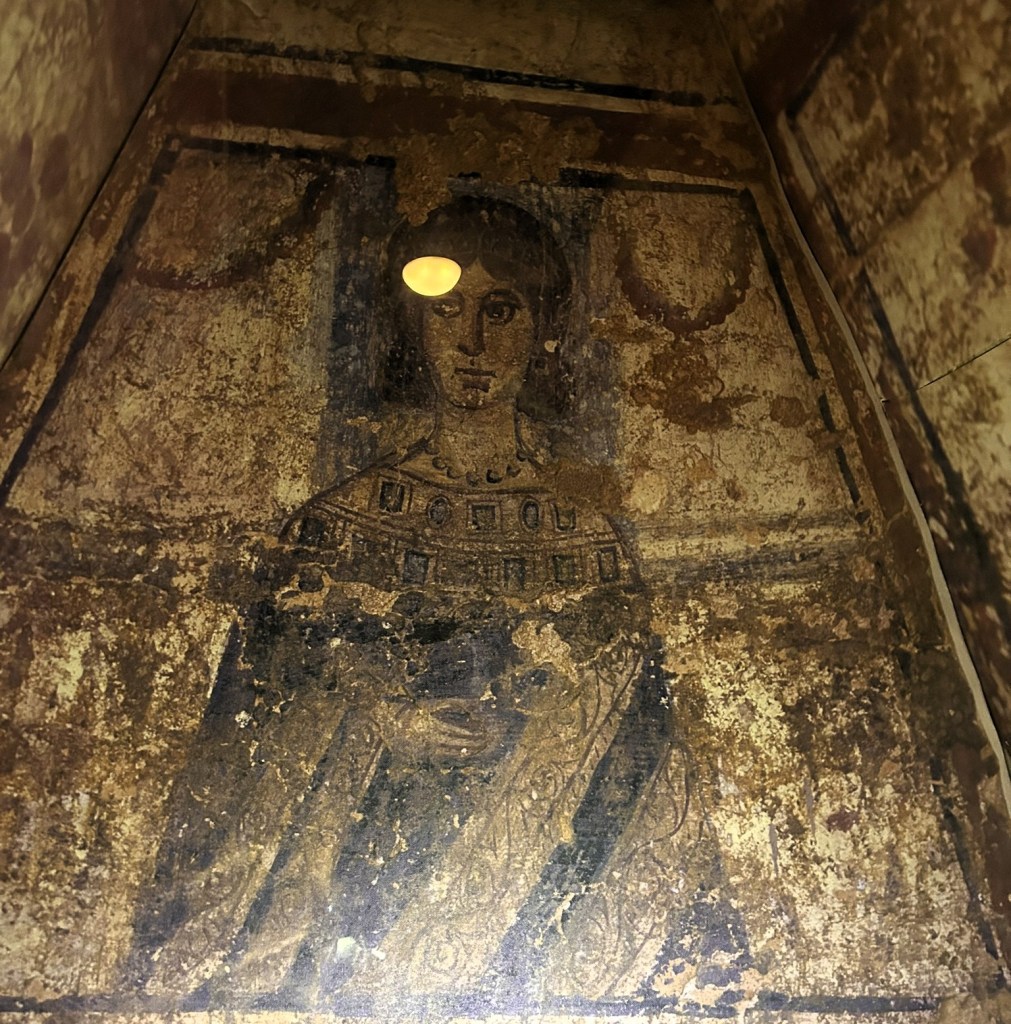 Ancient mural of a woman with long hair and patterned clothing, partially obscured by wear and tear.