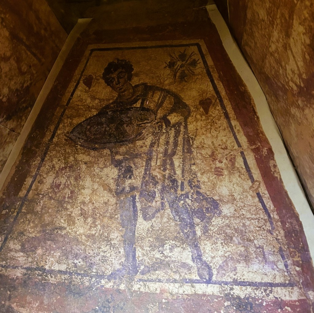 Ancient fresco depicting a male figure holding a shield, surrounded by fading colours and intricate patterns on a weathered wall.