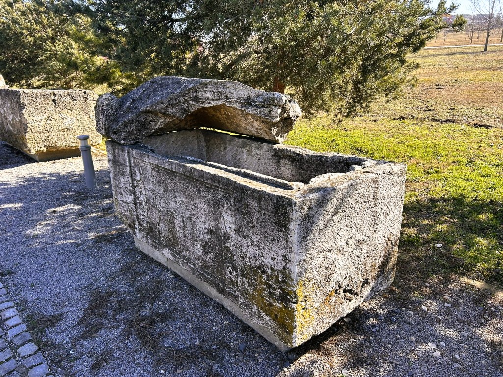 An ancient stone sarcophagus with a partially opened lid, situated in a grassy area surrounded by trees.
