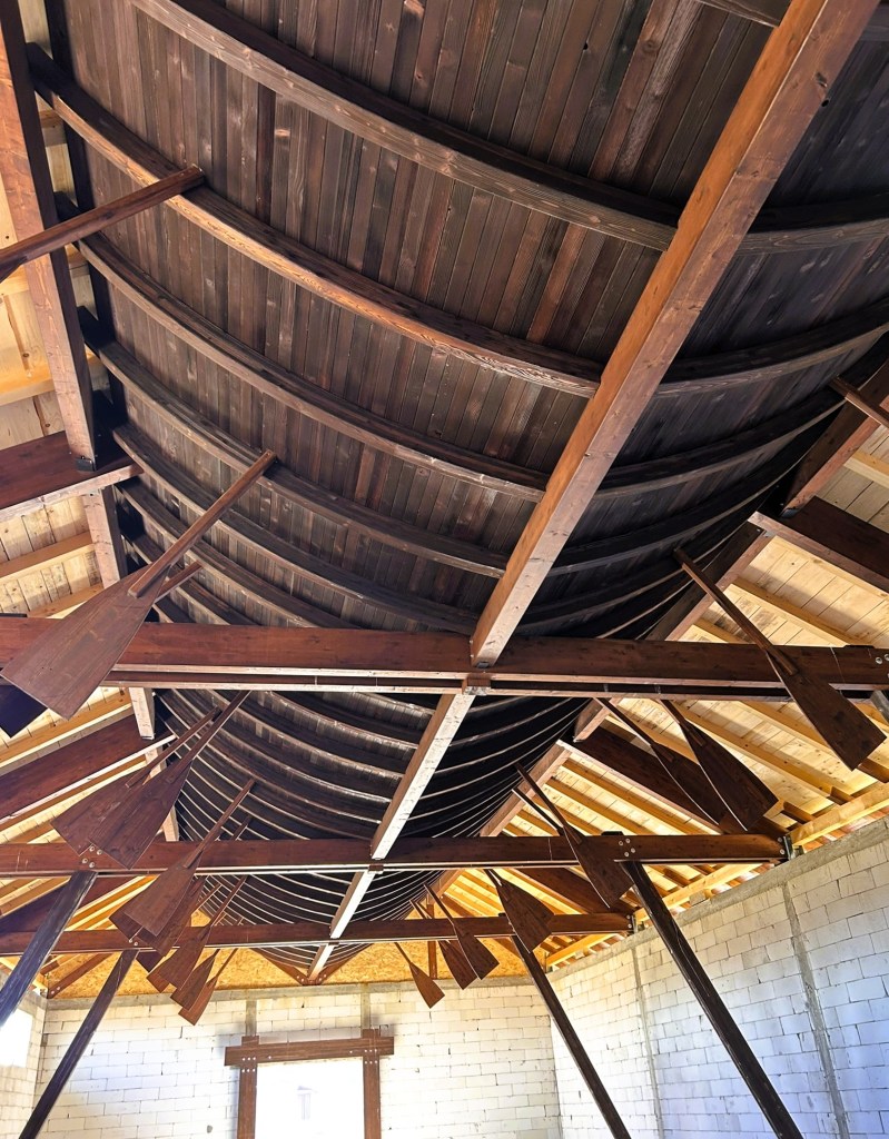 View of a wooden, curved ceiling structure supported by beams, with a rustic design featuring slanted wooden elements and a spacious interior.