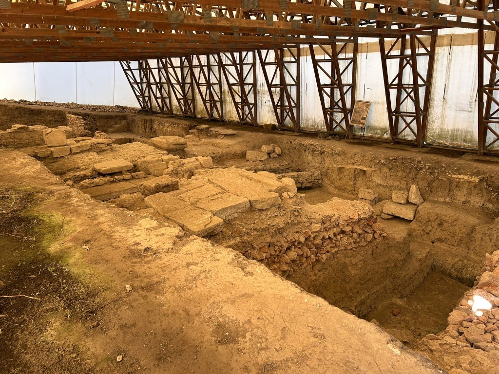 An archaeological excavation site showing exposed stone structures and layers of earth, covered by a wooden framework.