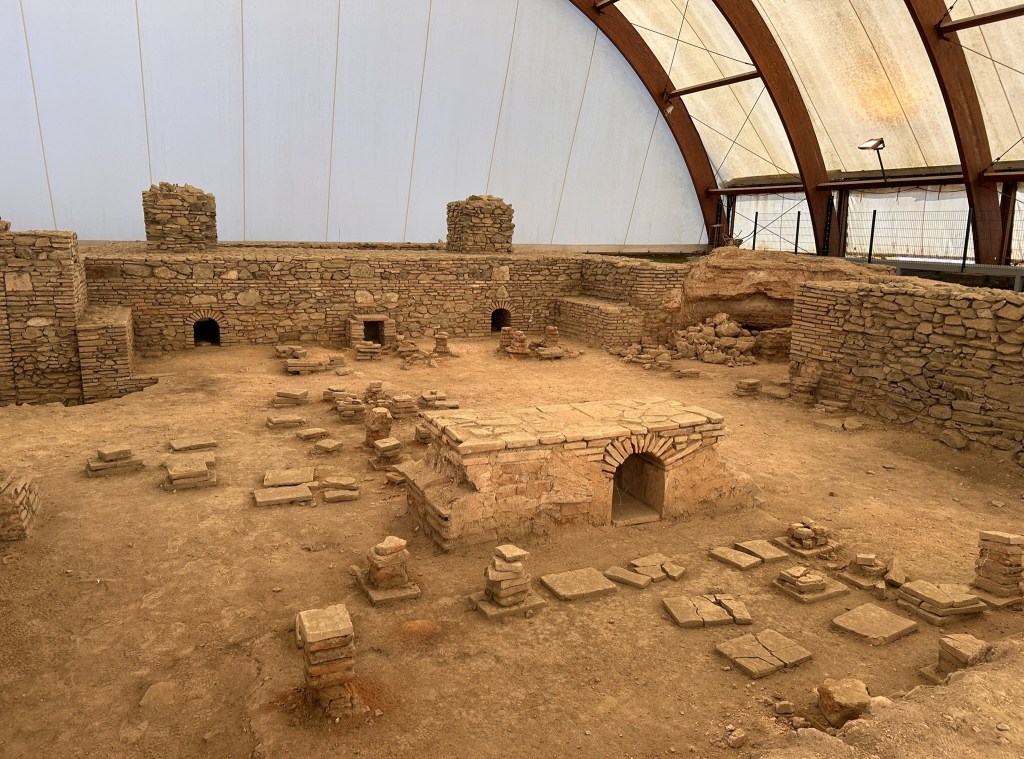 Interior view of an archaeological site under a protective structure, featuring stone walls, foundations, and scattered remnants of ancient structures.