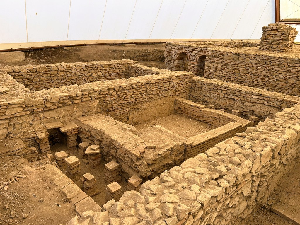 An archaeological site featuring stone ruins of an ancient building, with visible outline of walls, bricks, and stone blocks, covered by a protective structure.