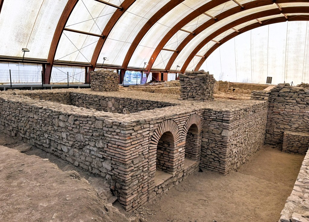 Excavated stone ruins of an ancient building featuring arched openings, housed under a large protective structure.