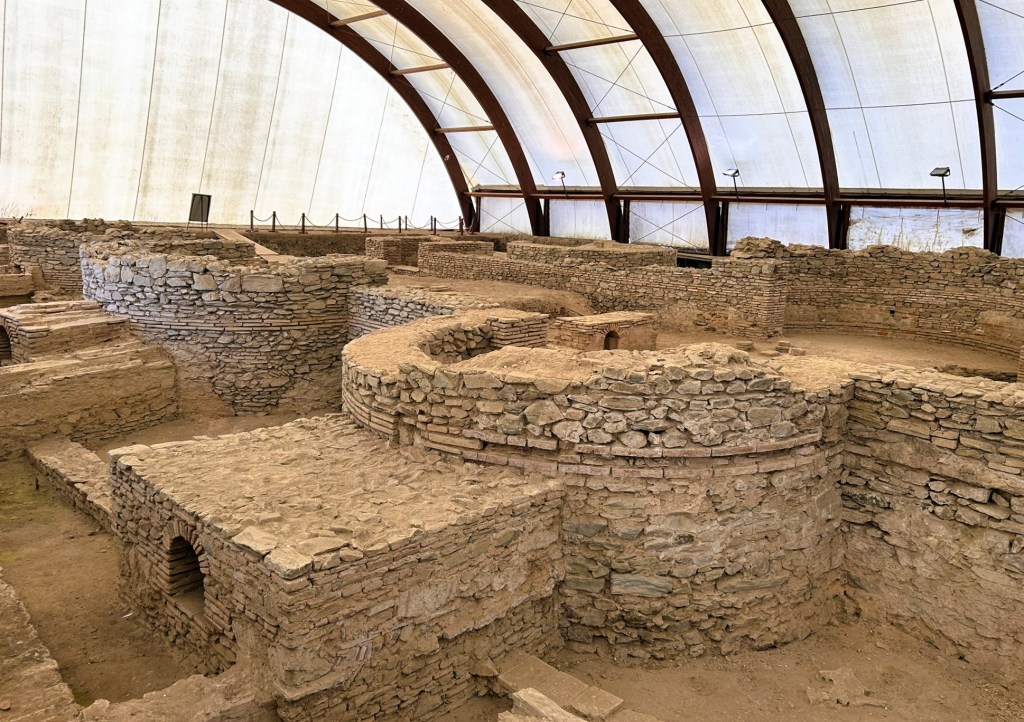 Excavated archaeological site featuring circular and rectangular stone structures under a protective canopy.