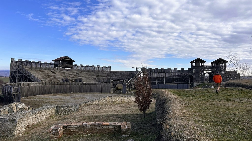 An ancient wooden fort with an arena, featuring stone ruins and a figure walking on grass, under a cloudy blue sky.