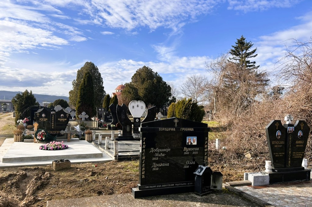 A peaceful cemetery scene featuring several gravestones, some adorned with flowers, surrounded by trees under a partly cloudy sky.