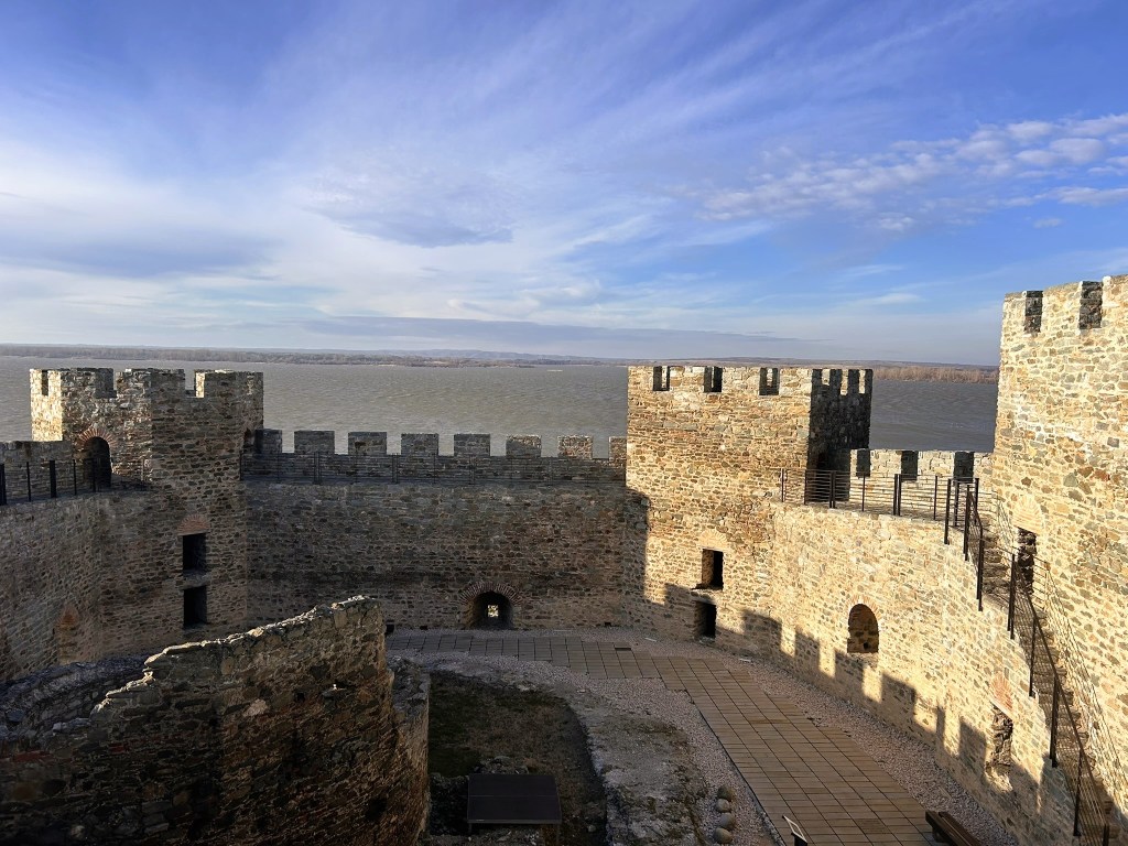 View of a medieval stone castle with battlements, overlooking a calm body of water under a partly cloudy sky.