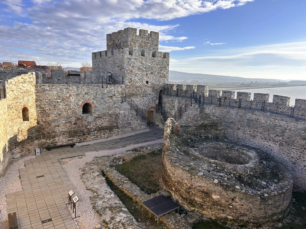 A view of a historic stone castle with a tall tower, surrounded by ancient walls and a grassy courtyard, under a partly cloudy sky.