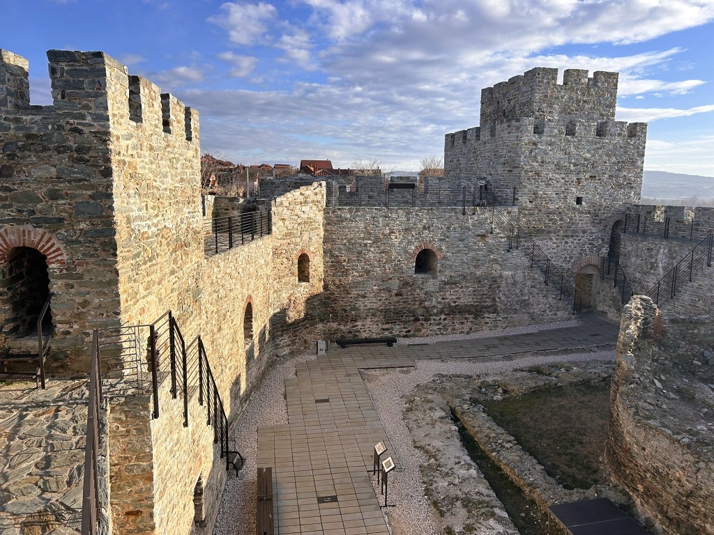 An aerial view of an ancient stone castle with crenellated walls and a circular tower, surrounded by pathways and grassy areas under a cloudy sky.