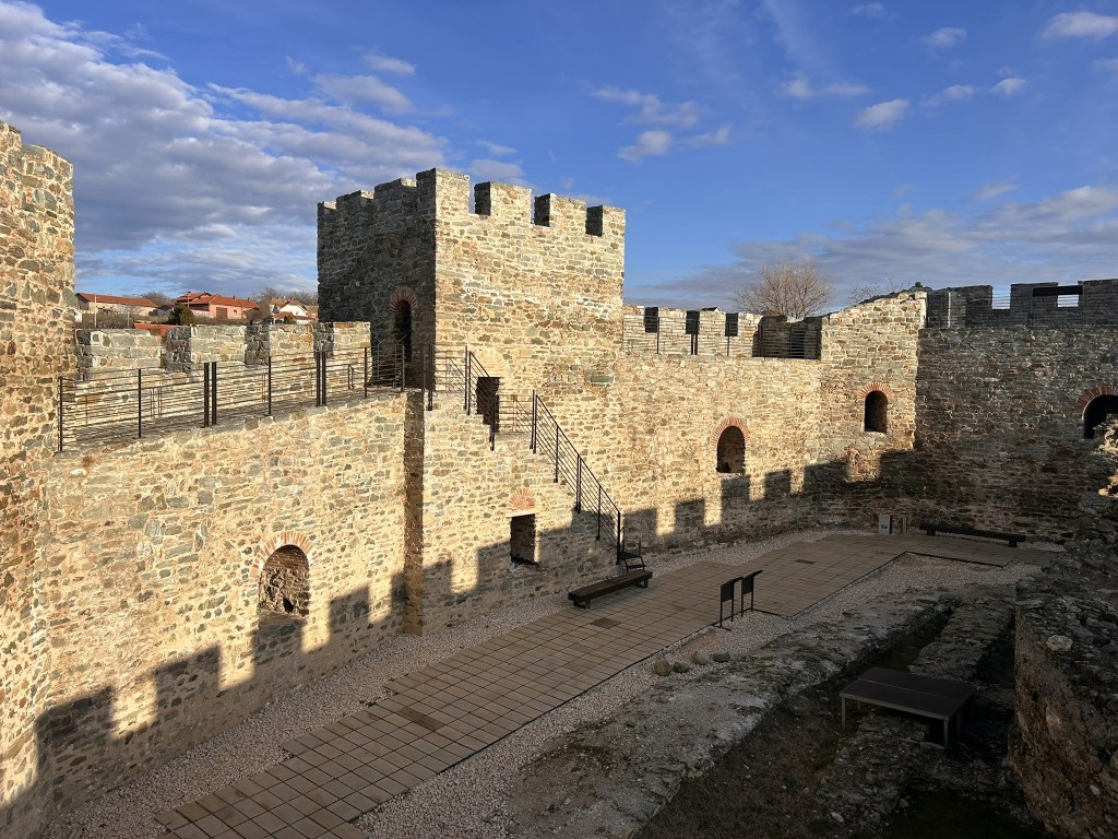Ruins of a stone castle with towers and a courtyard, featuring a stairway and benches, set against a cloudy sky.