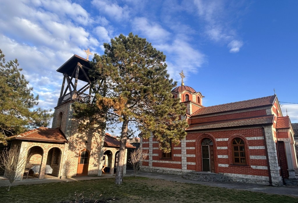 A brick and stone church building with a dome and cross, accompanied by a bell tower and trees, under a partly cloudy sky.