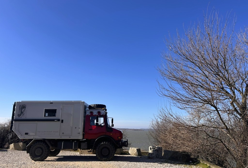 A red off-road vehicle parked on a gravel area by a body of water, with a bright blue sky and leafless trees in the background.