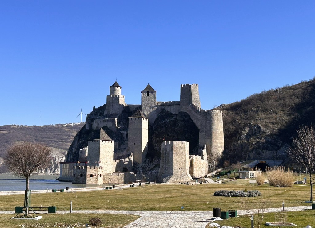 A medieval castle situated on a cliff by a river, featuring multiple towers and fortifications against a clear blue sky.