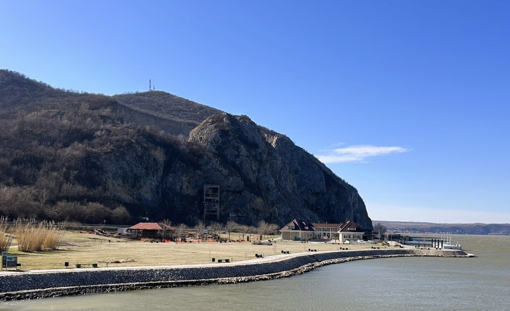 Scenic view of a riverside area with a large rocky hill in the background, featuring a house and seating area along the water's edge under a clear blue sky.