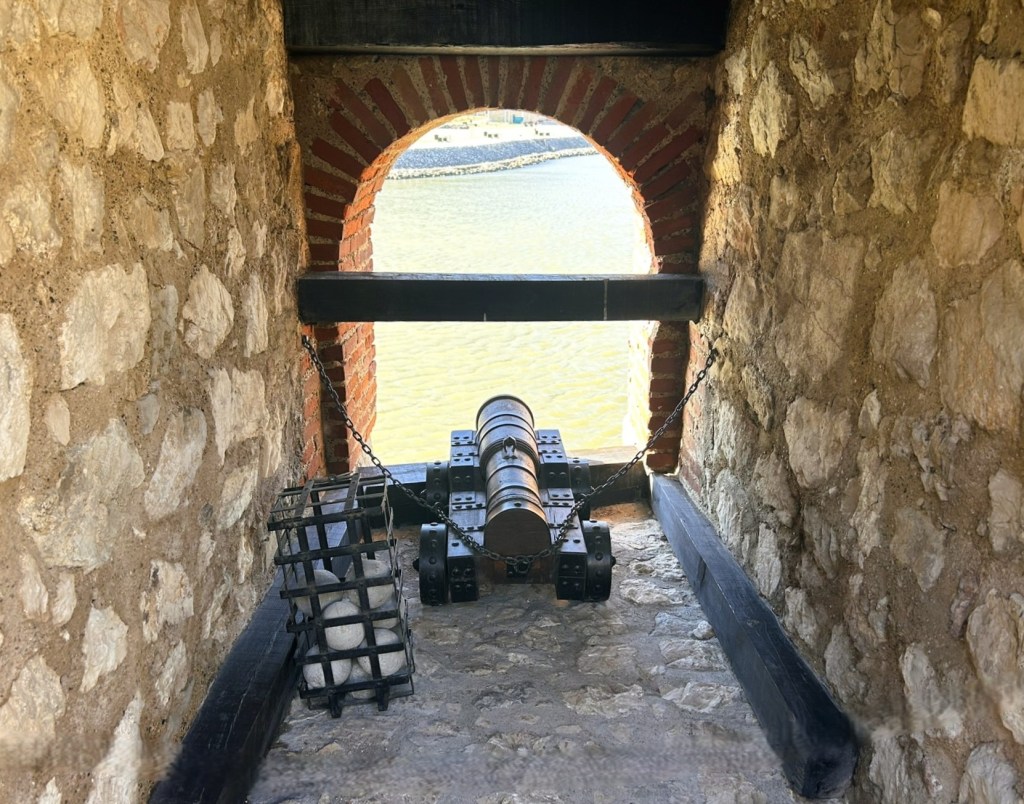 View of a historical cannon positioned behind a stone wall, with a wooden structure and a basket of cannonballs nearby, overlooking a water body.