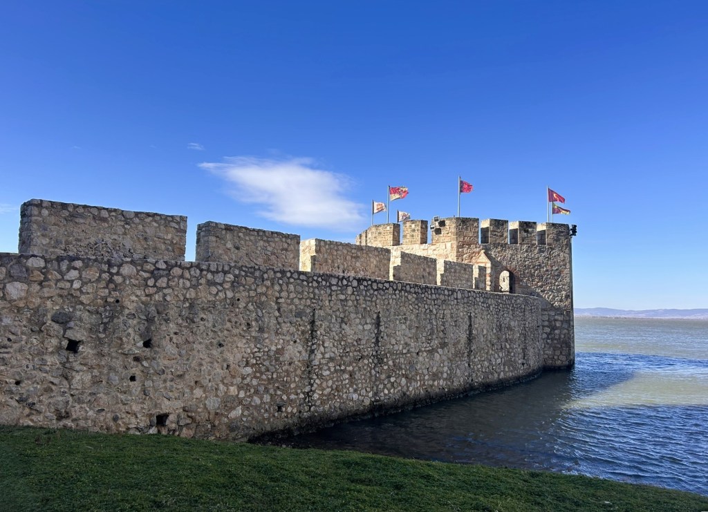 A stone castle with crenellated walls and flags on top, situated beside a body of water under a clear blue sky.