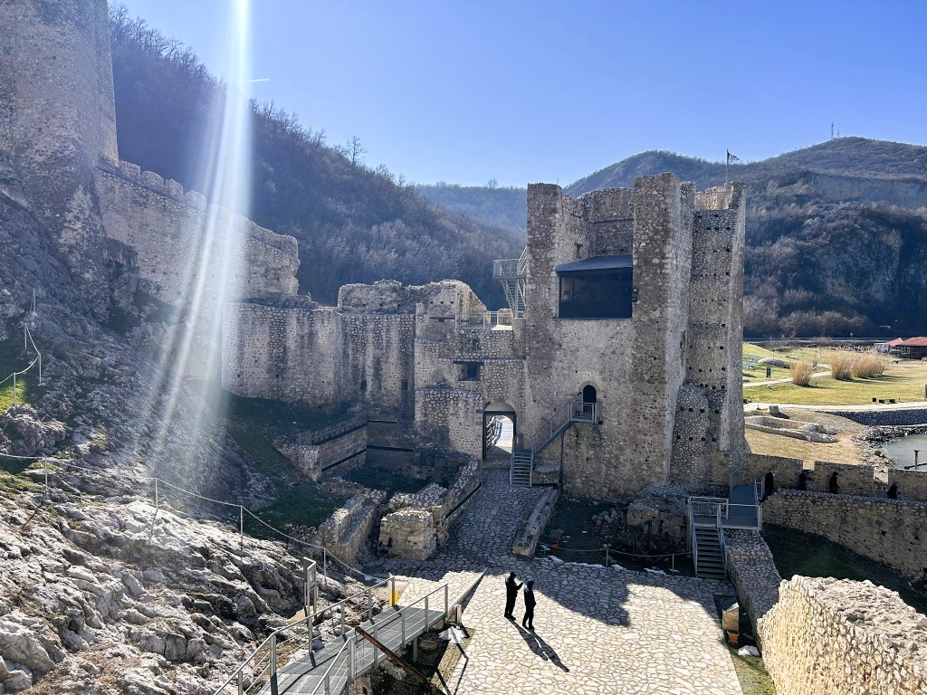 View of a historic castle with stone walls and towers, surrounded by rocky terrain and hills, under a clear blue sky. Two people are walking along a stone pathway in front of the castle.