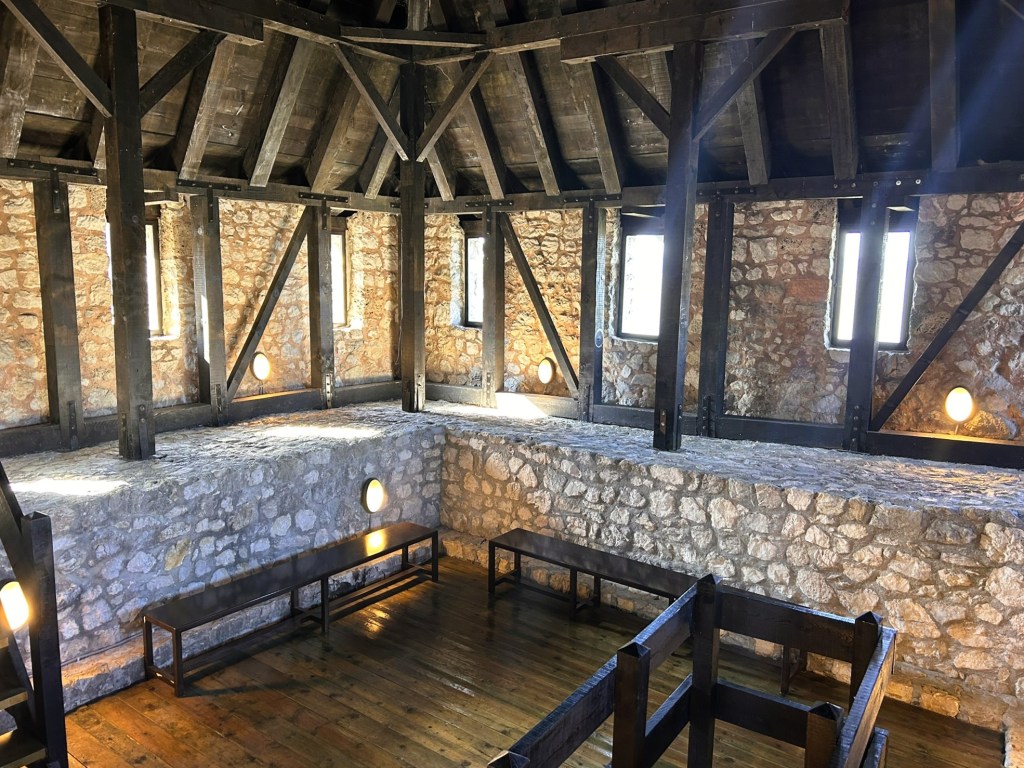 Interior view of a rustic stone room with wooden beams and benches, featuring natural light from small windows.