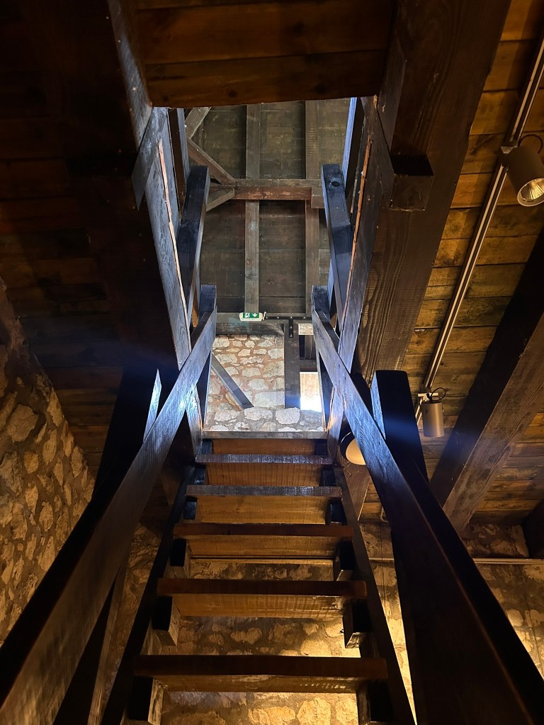View of wooden stairs leading up to a loft, with exposed beams and stone walls in the background.