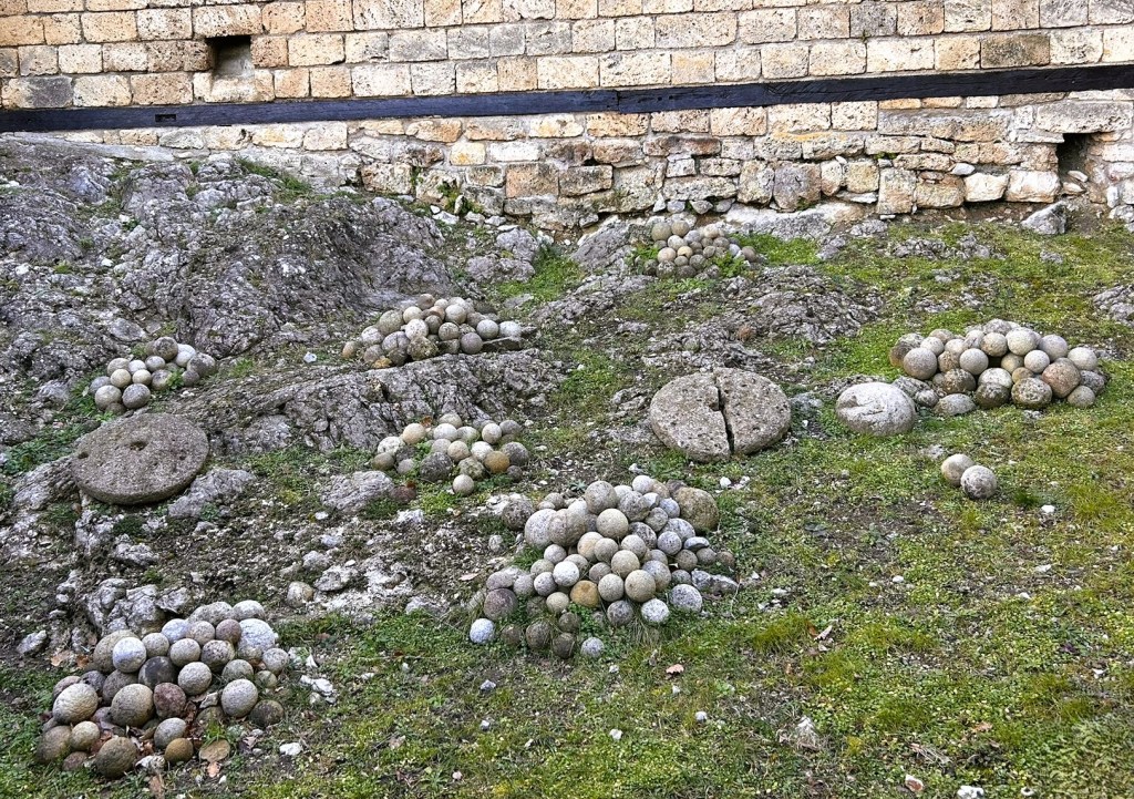 A collection of rounded stone balls of various sizes arranged in groups on a rocky surface, with a brick wall in the background.