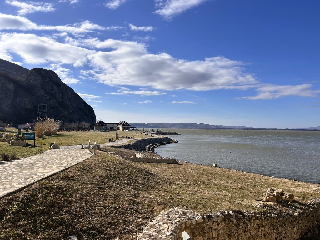Scenic view of a lake with a rocky shoreline and a pathway, framed by a mountain and a blue sky with clouds.