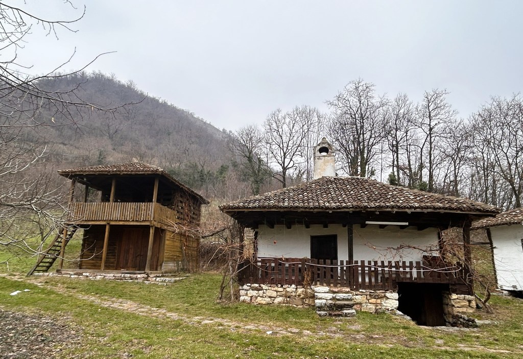 A picturesque scene of two traditional rural houses, one with a wooden balcony and the other featuring a stone foundation and a tiled roof, set against a backdrop of bare trees and a cloudy sky.