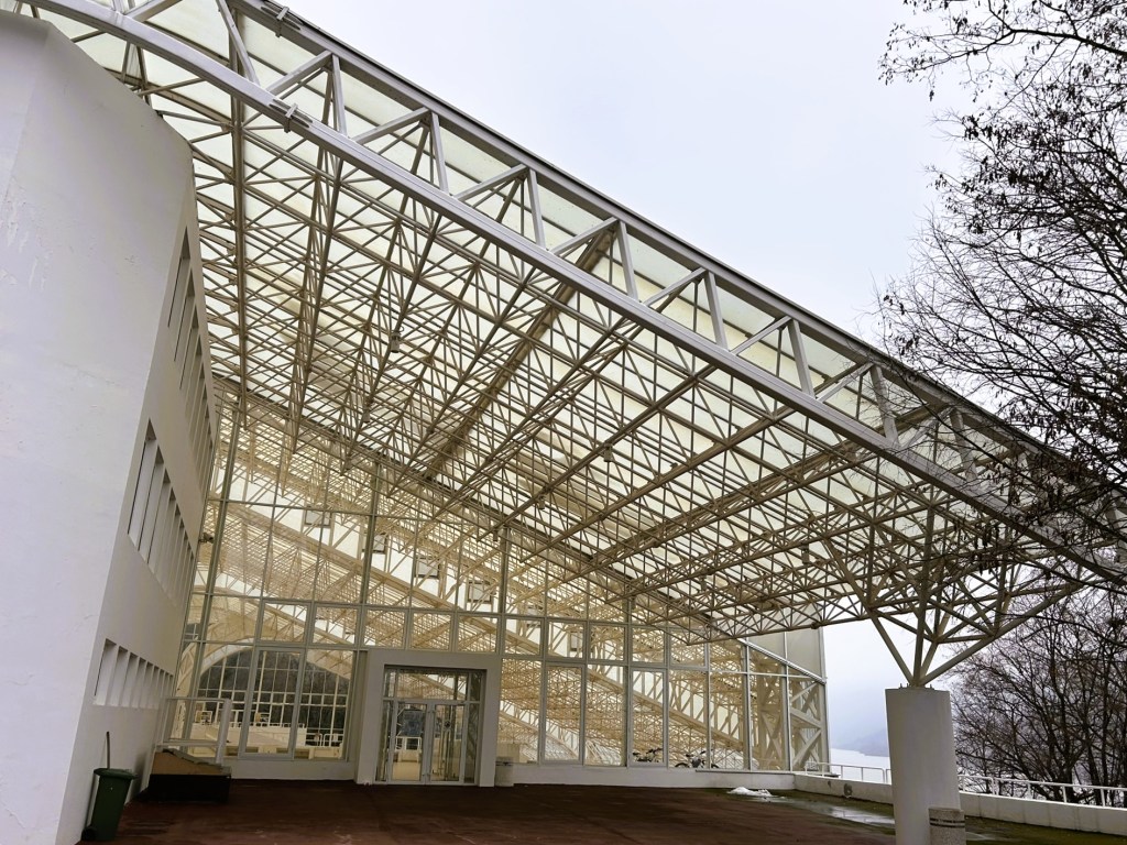 A modern architectural structure featuring a large glass roof supported by a white metal framework, with an entrance visible at the centre and trees in the background.
