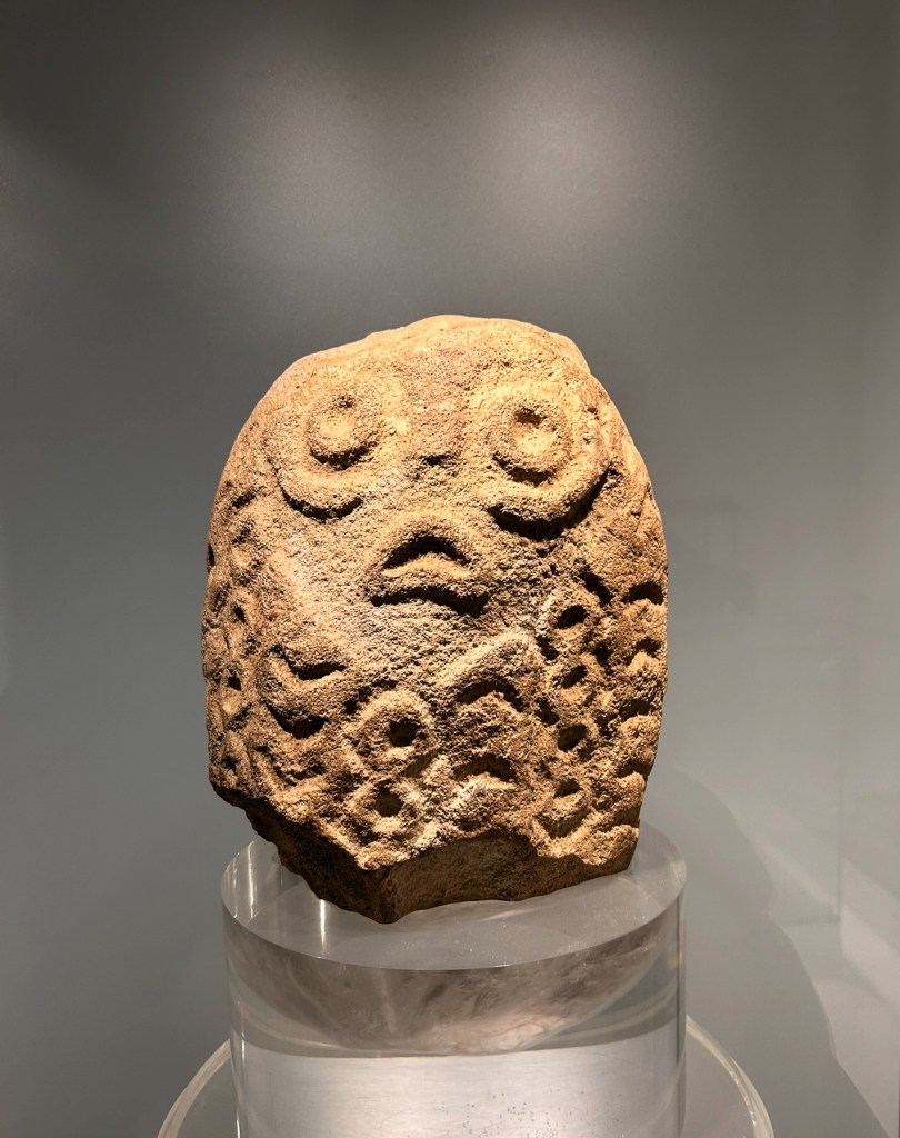 A stone sculpture featuring a stylised face with large eyes and an open mouth, exhibiting intricate carvings on its surface, displayed on a transparent pedestal.