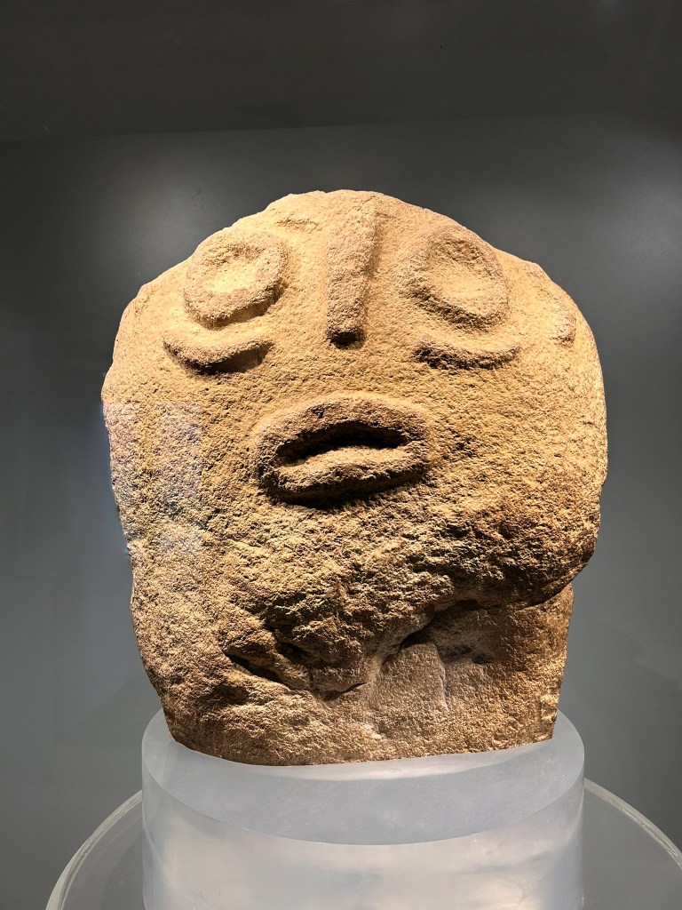 A stone sculpture resembling a face, featuring prominent eyes, a flat nose, and a slightly open mouth, displayed on a podium in a museum setting.