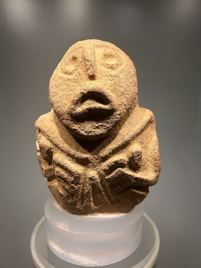 A stone sculpture depicting a humanoid figure with exaggerated facial features and outstretched arms, displayed on a clear pedestal.