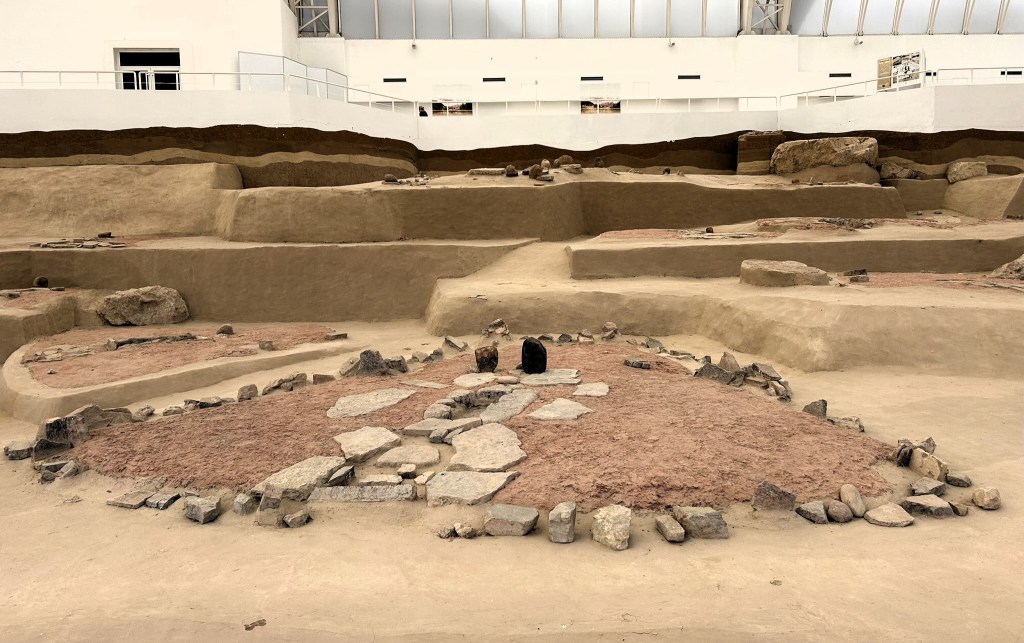 An archaeological site featuring layered earth mounds and scattered stones, showcasing remnants of ancient structures.