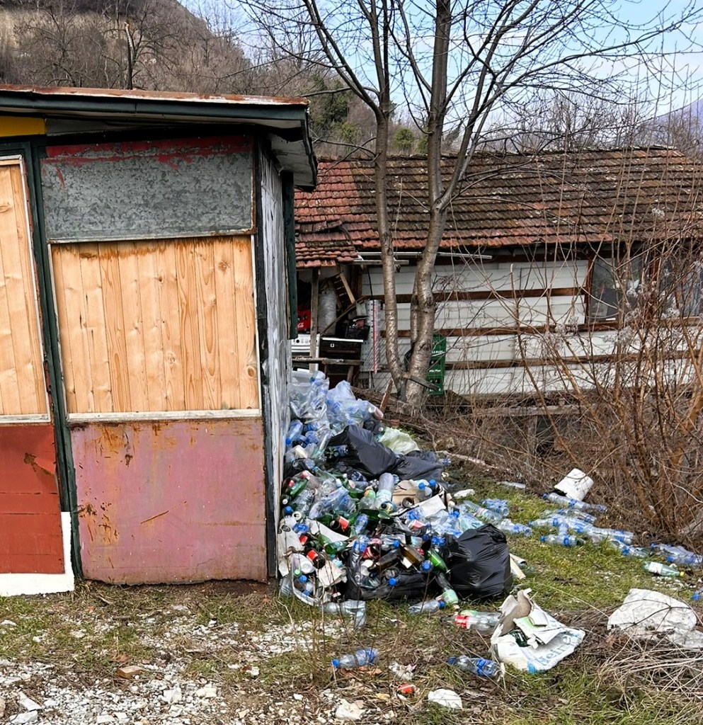 A rundown shed beside a large pile of discarded plastic bottles and garbage, surrounded by bare trees and a hilly background.