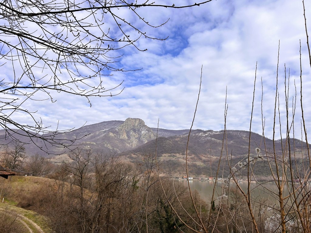A scenic view of a mountainous landscape with bare trees in the foreground, under a cloudy sky.