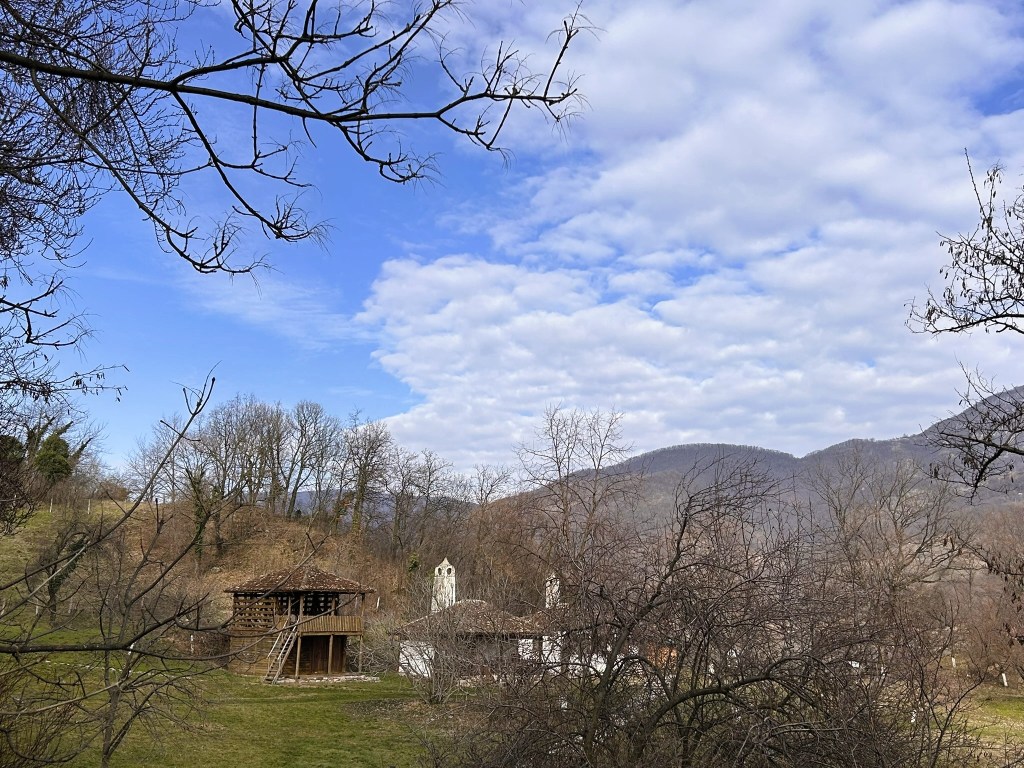 A scenic view of a rural landscape featuring a wooden structure and a traditional building, surrounded by leafless trees and rolling hills under a partly cloudy sky.