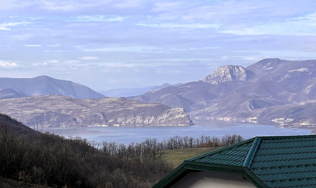 A scenic view of a lake surrounded by mountains, with a distant coastline visible. The foreground features a green roof, blending with the natural landscape.