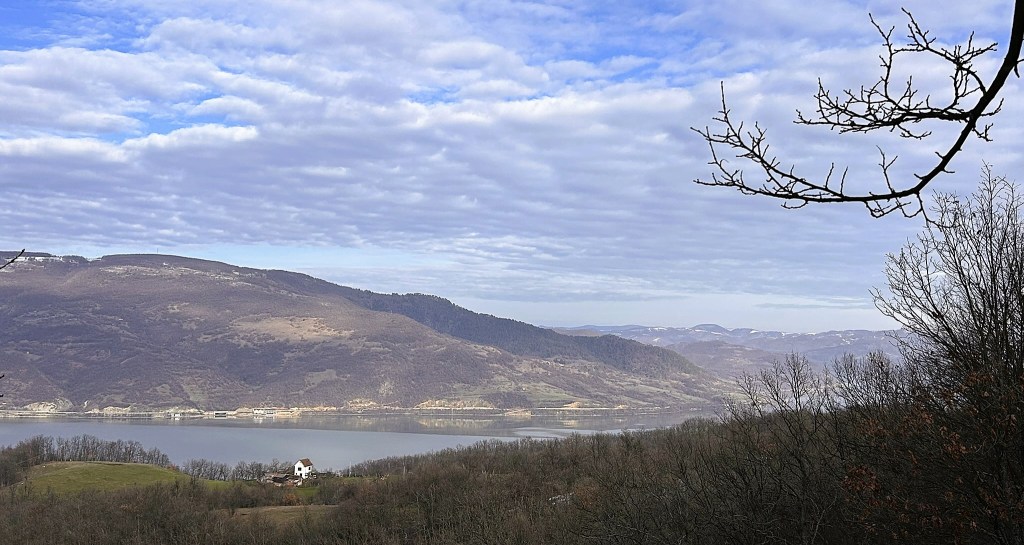 A scenic view of a mountainous landscape with a calm lake in the foreground, featuring a small white house and bare trees in the foreground against a backdrop of cloudy skies.