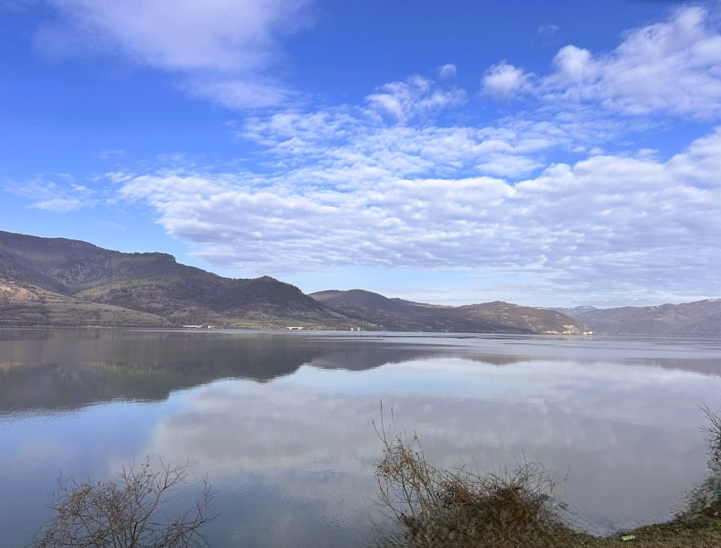 A tranquil landscape featuring a calm lake reflecting the blue sky and fluffy clouds, surrounded by rolling hills and mountains.