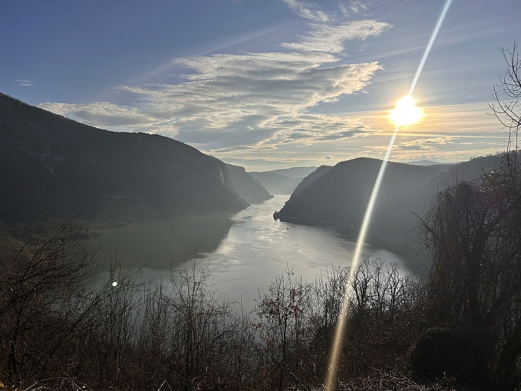 A serene view of a river winding between two mountains at sunset, with a bright sun casting rays over the landscape and soft clouds in the sky.