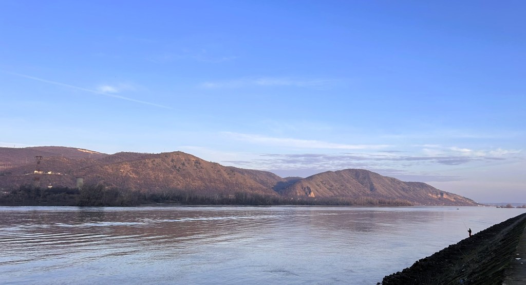 A scenic view of a river surrounded by hills under a clear blue sky, with a lone fisherman standing on the riverbank.