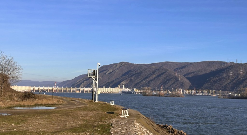 A scenic view of a river landscape with mountains in the background, featuring a dam structure along the water's edge and a clear blue sky.