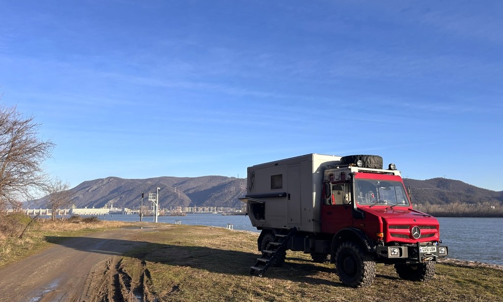 A red off-road camper truck parked by a riverbank, with mountains in the background and a clear blue sky.