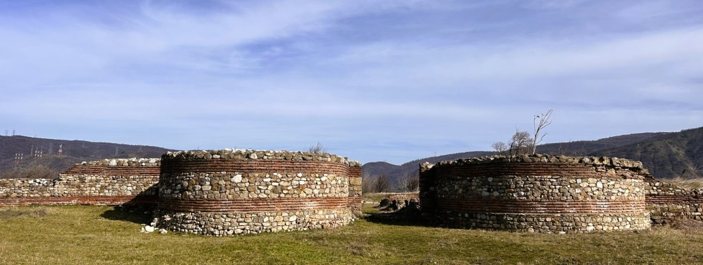 Ancient stone ruins with circular structures, surrounded by grassy land and distant hills under a blue sky.