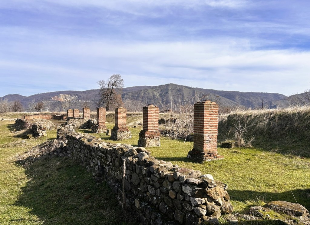 Ruins of ancient brick pillars and stone walls in a grassy landscape with hills in the background.