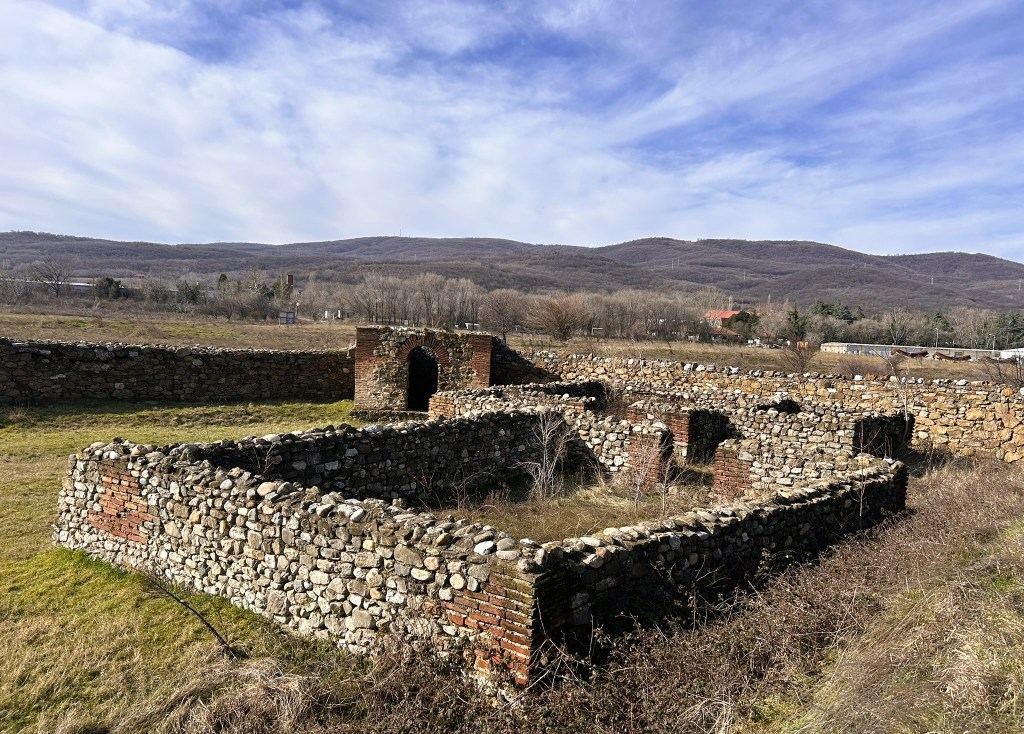 Ruins of an old stone building surrounded by vegetation and mountains in the background under a cloudy sky.