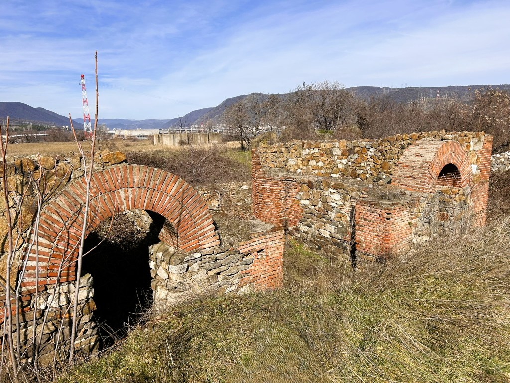Ruins of a stone structure with brick arches in a grassy field, surrounded by hills and a clear sky.