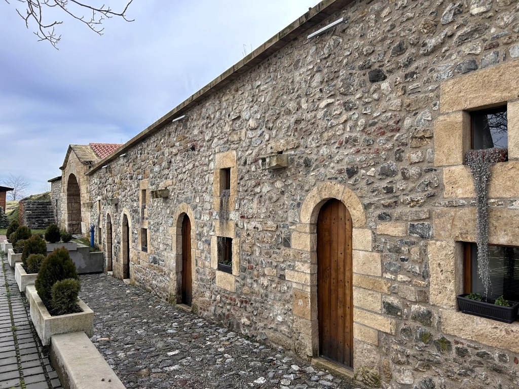 A stone building featuring multiple arched doorways and windows, with a cobblestone pathway and small shrubs in planters alongside the structure.