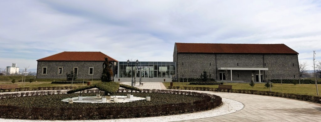 A landscaped park featuring a circular garden with a fountain, surrounded by stone buildings with red roofs and glass elements, under a cloudy sky.