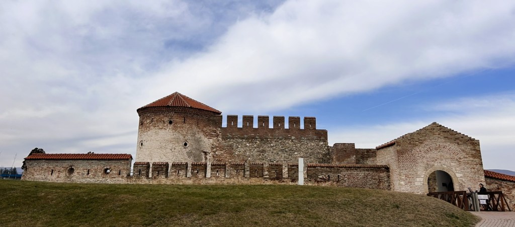 A historic stone fort with a red-tiled roof, featuring battlements and a circular tower, set against a cloudy sky.