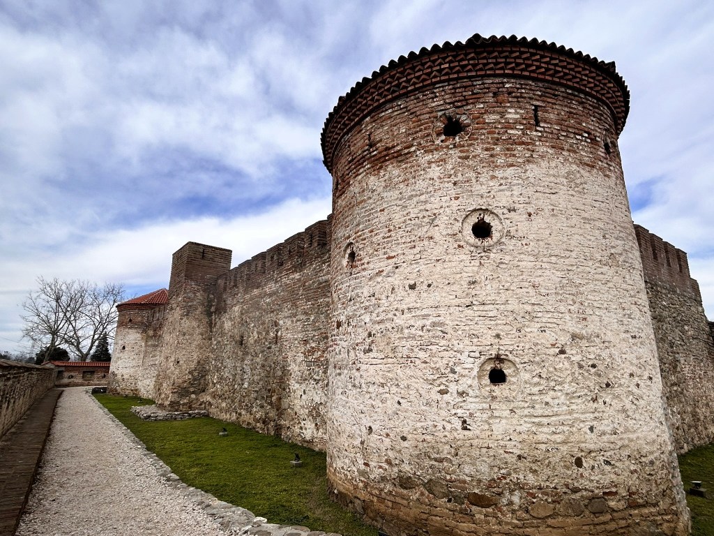 A stone castle wall with two round towers and a pathway beside it, under a cloudy sky.